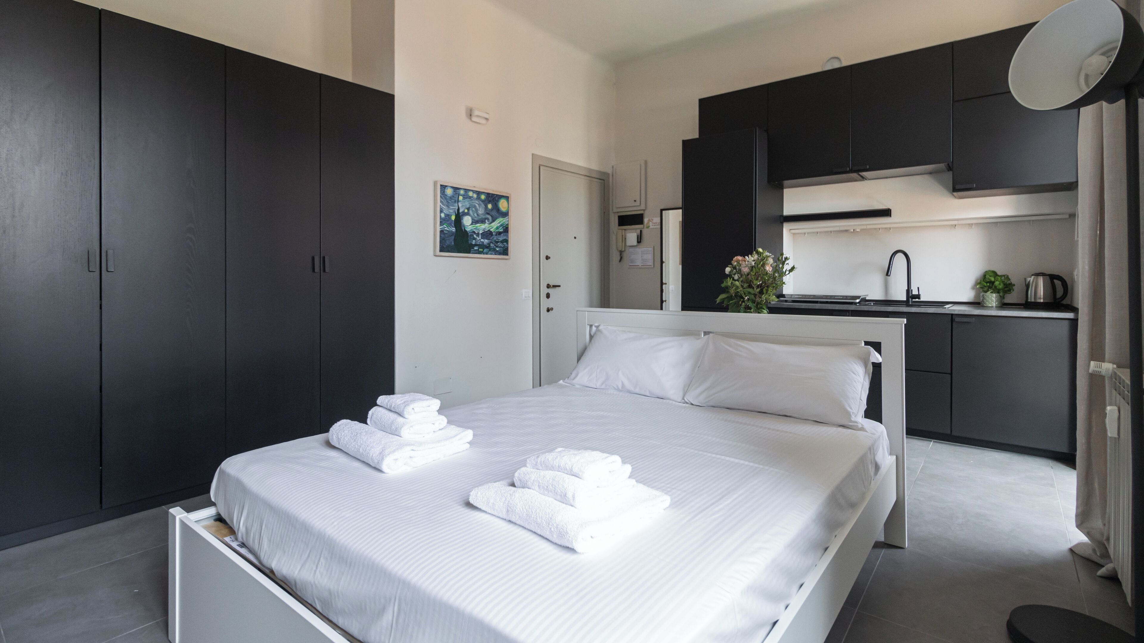 Habitació d'hotel moderna amb un llit ben fet, una petita zona de cuina i armaris de tons foscos.