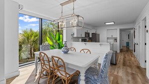A modern kitchen with white cabinetry, a dining area with wicker chairs, and a large window offering a view of greenery.