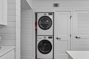 A laundry room with two stacked washers and dryers, white walls, and a door with a handle. - Shipwatch 206A - Coastal Luxury! Ocean View! (Isle of Palms)