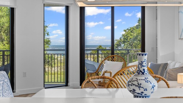 A room with a large window overlooking a beach, a wicker chair, and a white table with a blue vase.