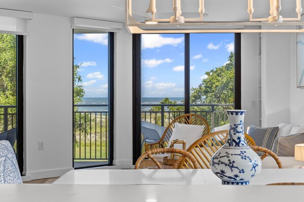 Una habitación con un ventanal grande con vista a una playa, una silla de mimbre y una mesa blanca con un florero azul.