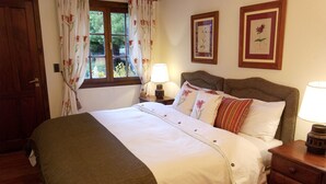 A neatly made bed with a green blanket, a wooden nightstand with a lamp, and two framed botanical prints on the wall. - Hosteria Helsingfors (Estancia Helsingfors)
