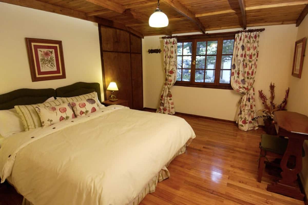 Een slaapkamer met een houten plafond, een bed met bloemenbedrukking, een nachtkastje met een lamp, een raam met bloemen gordijnen en een houten stoel.