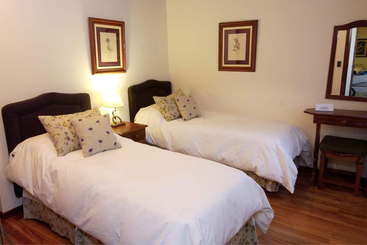 Een hotelkamer met twee enkele bedden, een nachtkastje met lamp, een houten bankje en een spiegel aan de muur.