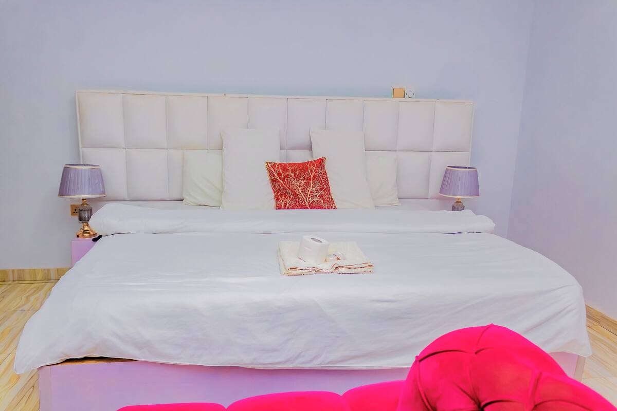 Una cama bien hecha con ropa blanca, una almohada decorativa roja y un par de toallas dobladas.