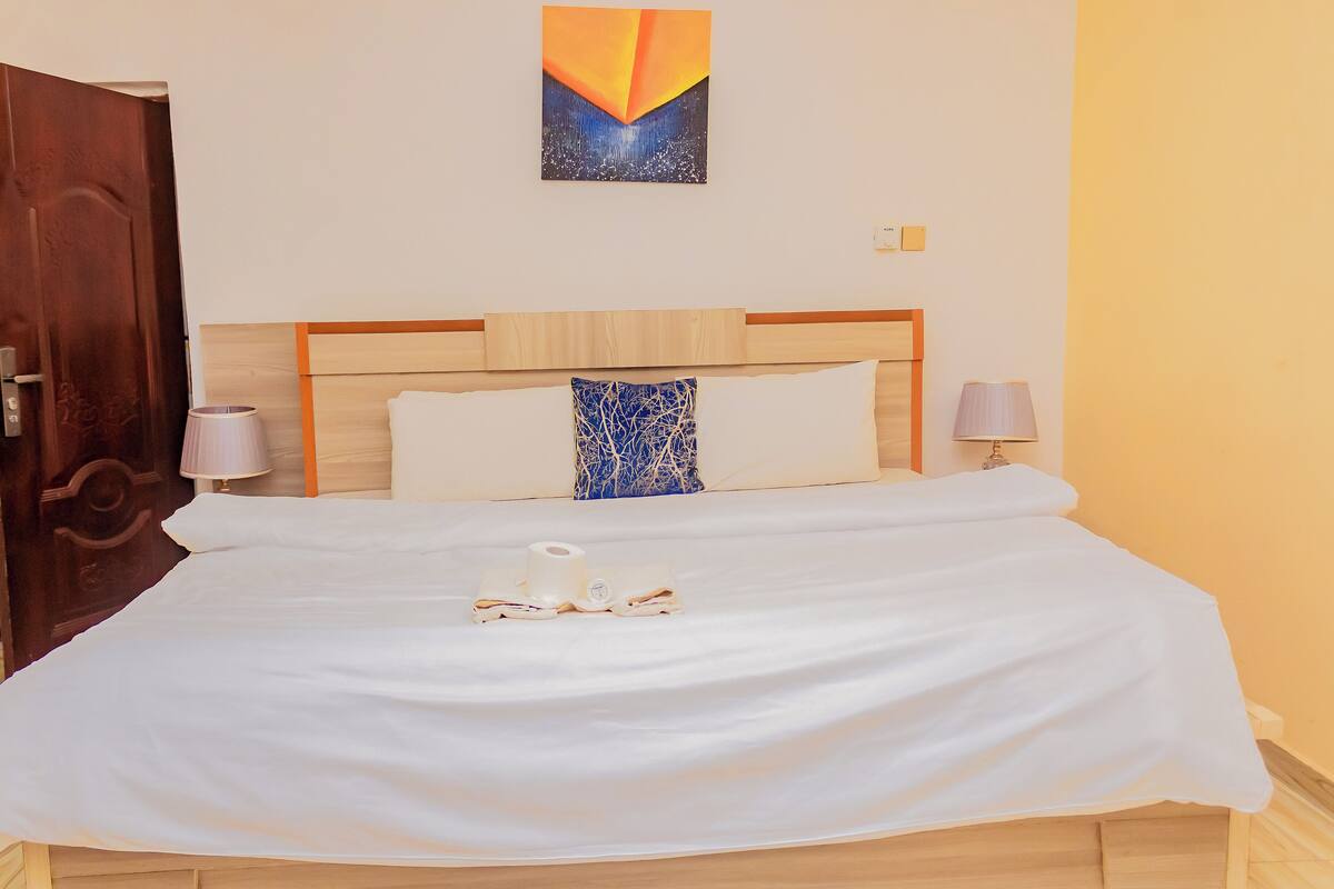 Una cama bien hecha con sábanas blancas, una almohada de diseño azul y un rollo de papel higiénico sobre la cama.