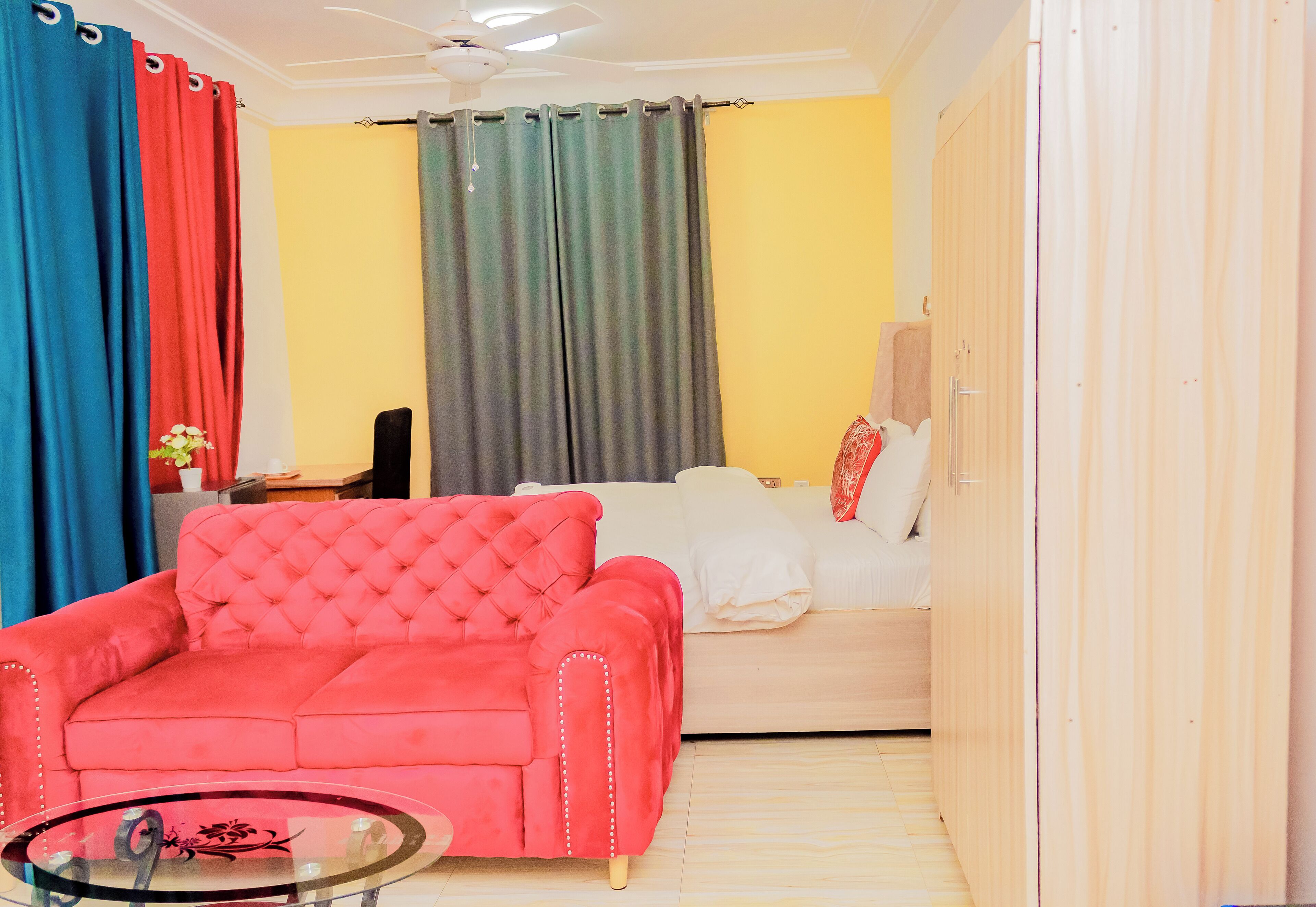 Una habitación con un sofá rojo, un sofá blanco y una cama. Se observa una mesa de vidrio al frente.