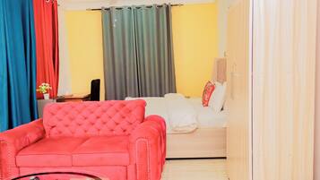 Una habitación con un sofá rojo, un sofá blanco y una cama. Se observa una mesa de vidrio al frente.