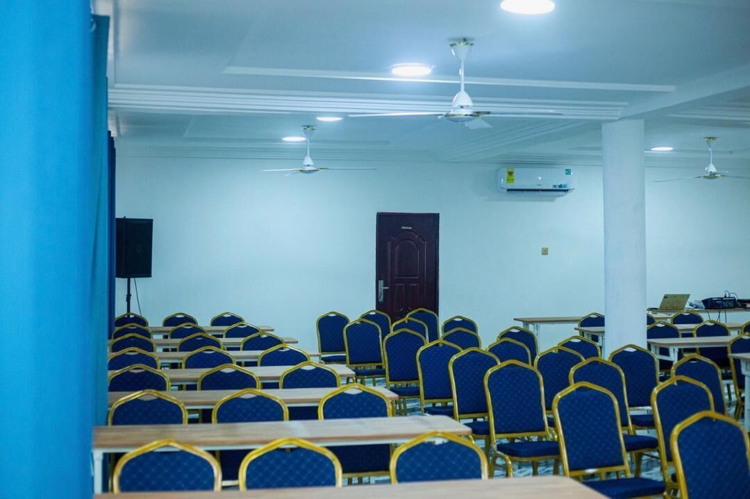 Un salón de conferencias con filas de sillas y mesas azules, ventiladores de techo y una puerta al fondo.