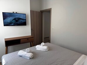 A hotel room with a bed, a TV mounted on the wall, a wooden desk, and a door leading to another room. - Massalia Suites Eolia (Corfu)