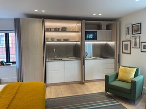 A compact living space with a kitchenette, a sofa with a yellow pillow, and a window with curtains. - Luxury Suite in Cadogan Square III (London)