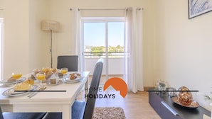 A dining area with a table set for breakfast, a view of the outdoors through a large window, and a modern interior design. - #320 Family Apartment in the City Center (Albufeira)