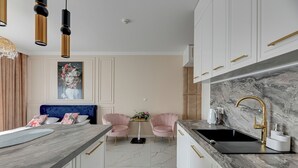 A modern kitchen with a marble backsplash, white cabinets, and gold handles. There is a black sink with a gold faucet, a marble countertop, and a small table with chairs. - RentPlanet - Studio Wałowa (Gdańsk)