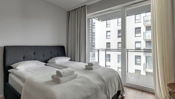 A modern bedroom with a large bed, grey curtains, and a view of a building through a glass door. - RentPlanet - Studio Wałowa (Gdańsk)
