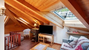 A cozy attic room with a sloped ceiling, wooden beams, a TV on a wooden stand, a sofa with decorative pillows, a small table, and a window overlooking a residential area.