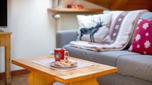 A cozy living room with a wooden table, a red mug, and cookies. A grey sofa with patterned pillows is in the background.