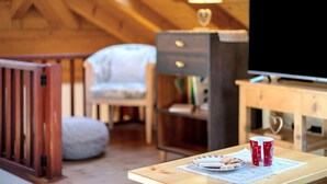 A cozy room with a wooden ceiling, a television on a wooden stand, a lamp, a chair, and a table with a mug and plate.