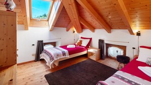 A bedroom with a sloped ceiling, wooden beams, a bed with a red blanket, a nightstand, a window with a view, and a small stool.