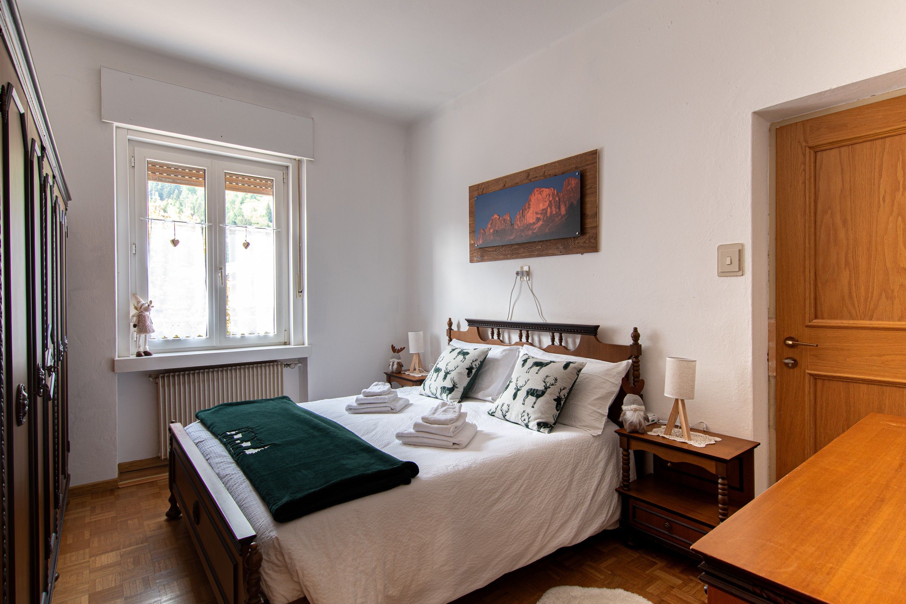 A bedroom with a bed, a window, a painting, a nightstand, and a wooden door.