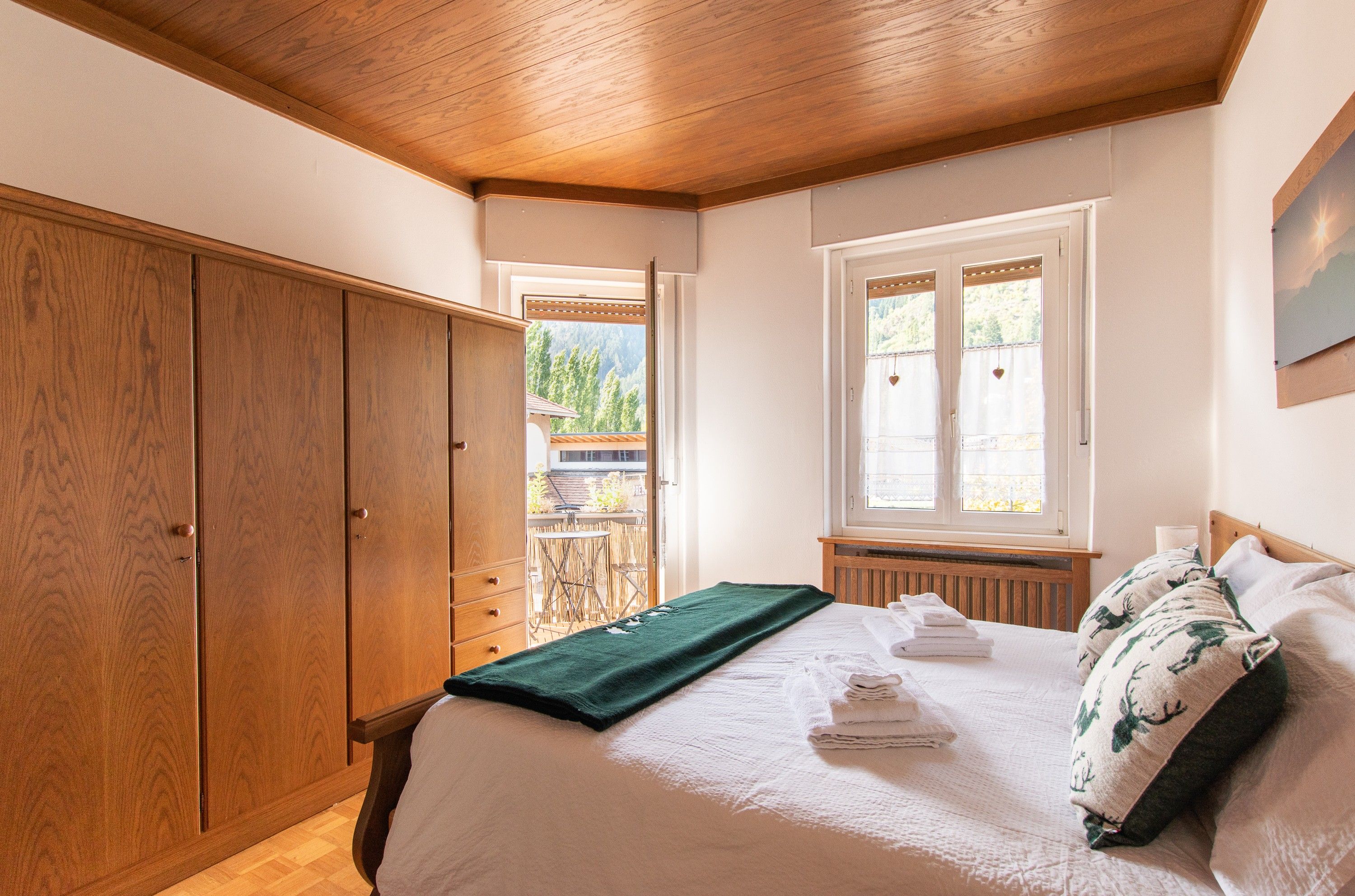 A bedroom with a wooden wardrobe, a bed with a green blanket, and a window with a view of greenery.