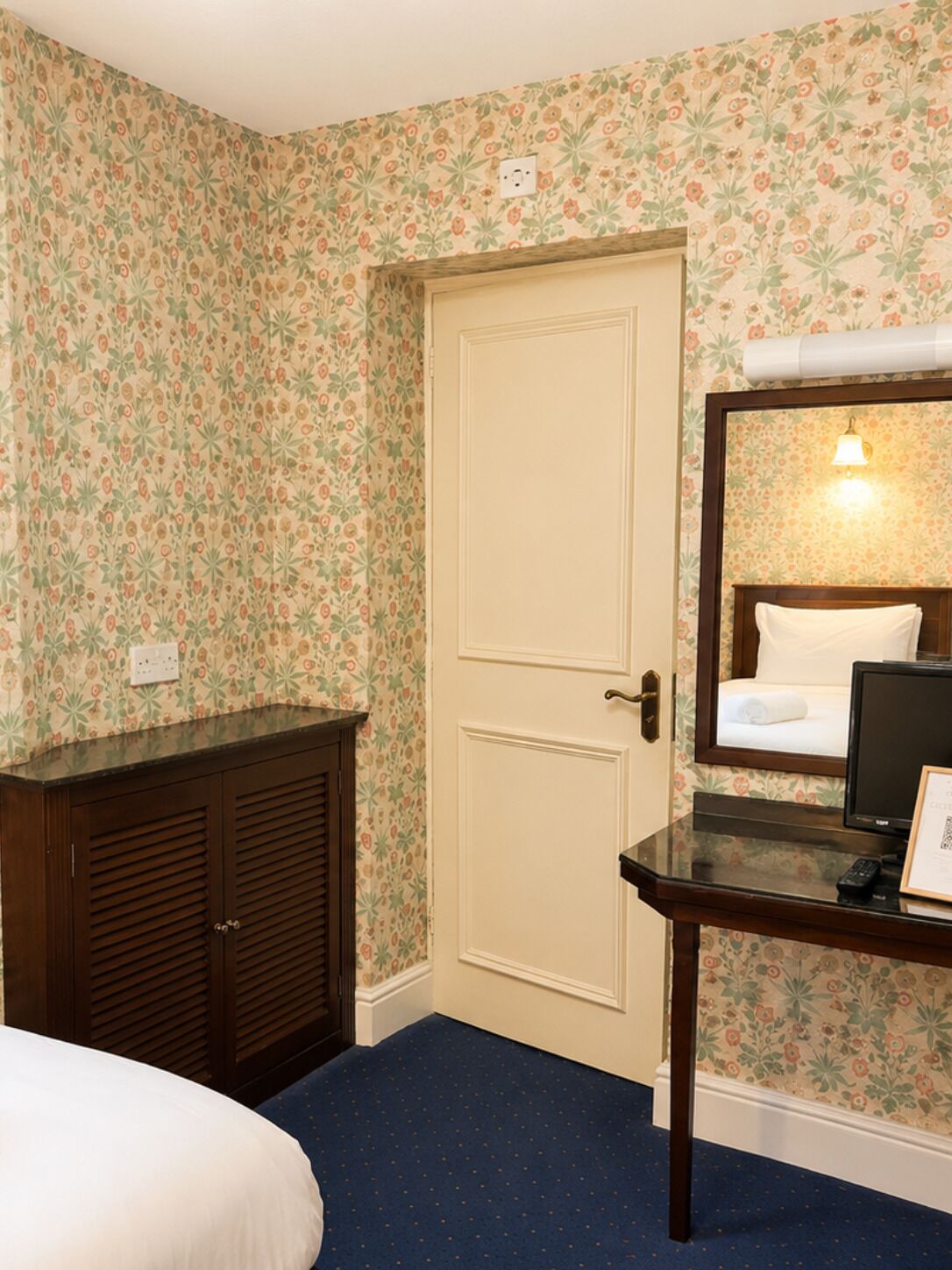 Ein Hotelzimmer mit floralem Wandpapier, einem Holzschrank, einem Bett mit weißer Daunendecke, einem Nachttisch mit Lampe und einem gerahmten Zertifikat.