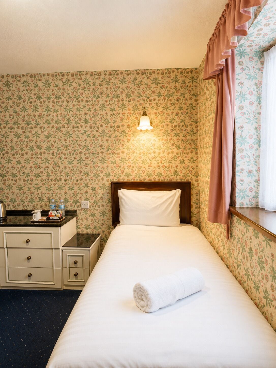 Een hotelkamer met een bed, een nachtkastje, een dressoir, een wandlamp en bloemenbehang.