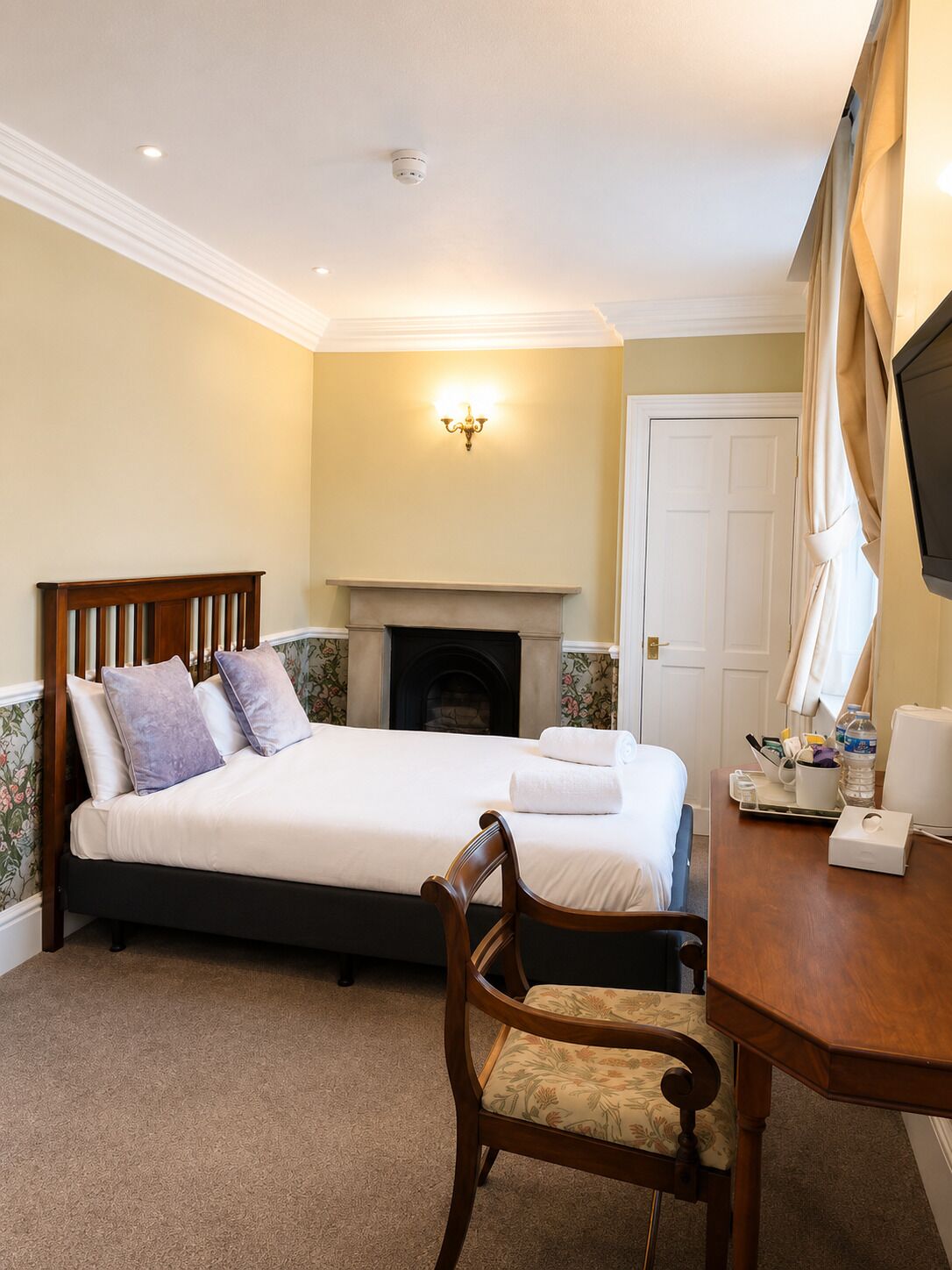 A hotel room with a bed, a chair, a desk with a lamp, a fireplace, and a television.