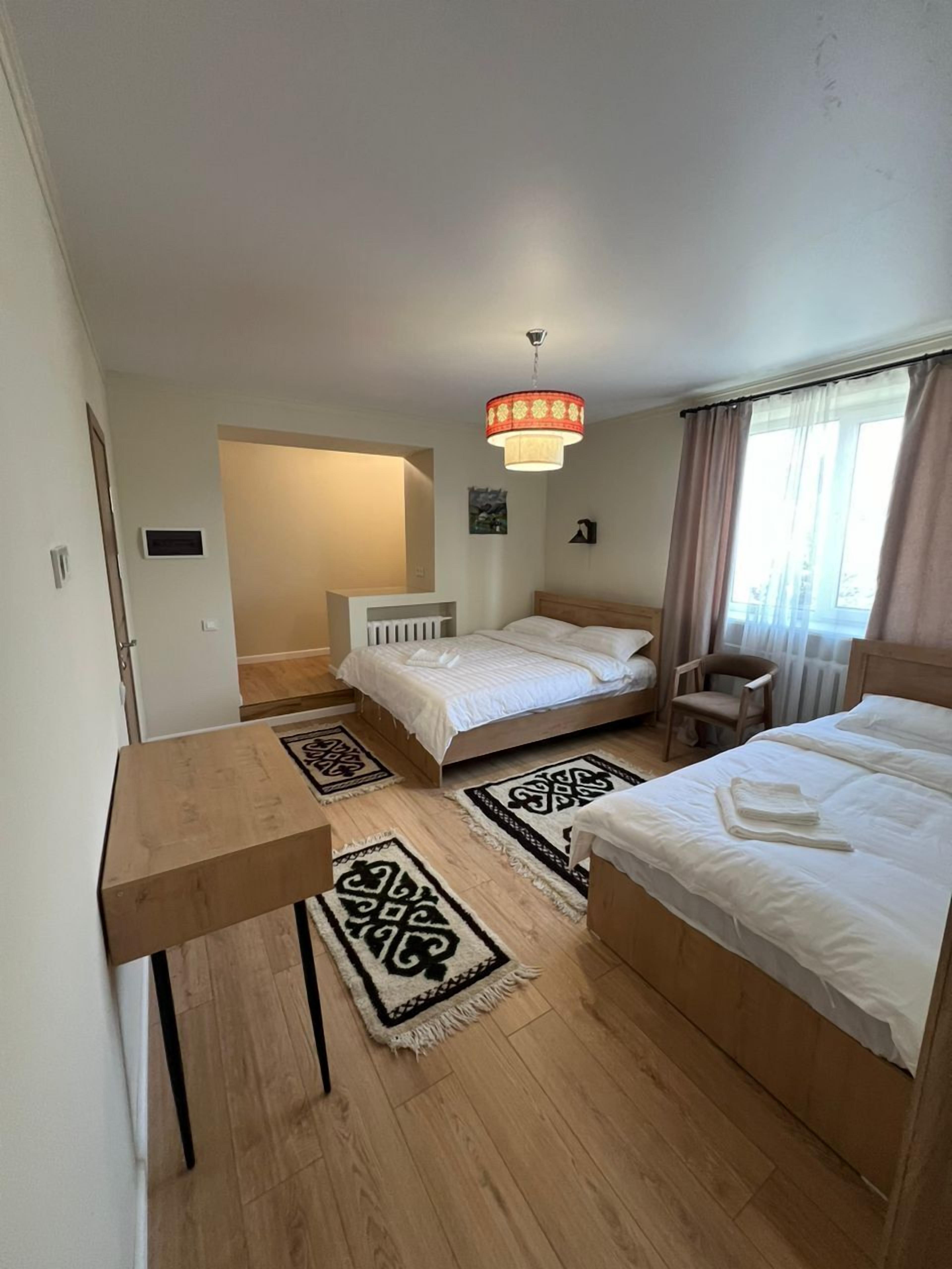 A hotel room with two beds, a wooden table, a chair, a radiator, and a large window with curtains.