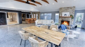 A spacious living area with a wooden dining table, wicker chairs, a stone fireplace, and a view of the outdoors. - Aqua Tides – Stylish Waterfront Escape in Busselton (Geographe)