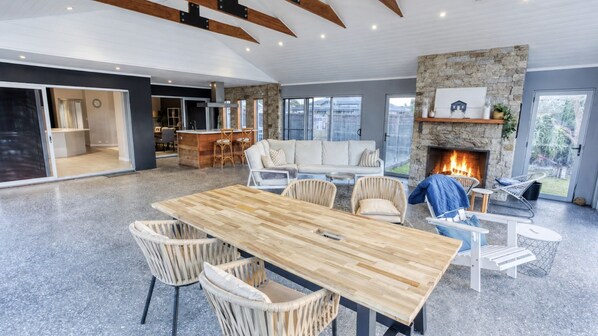 A spacious living area with a wooden dining table, wicker chairs, a stone fireplace, and a view of the outdoors. - Aqua Tides – Stylish Waterfront Escape in Busselton (Geographe)