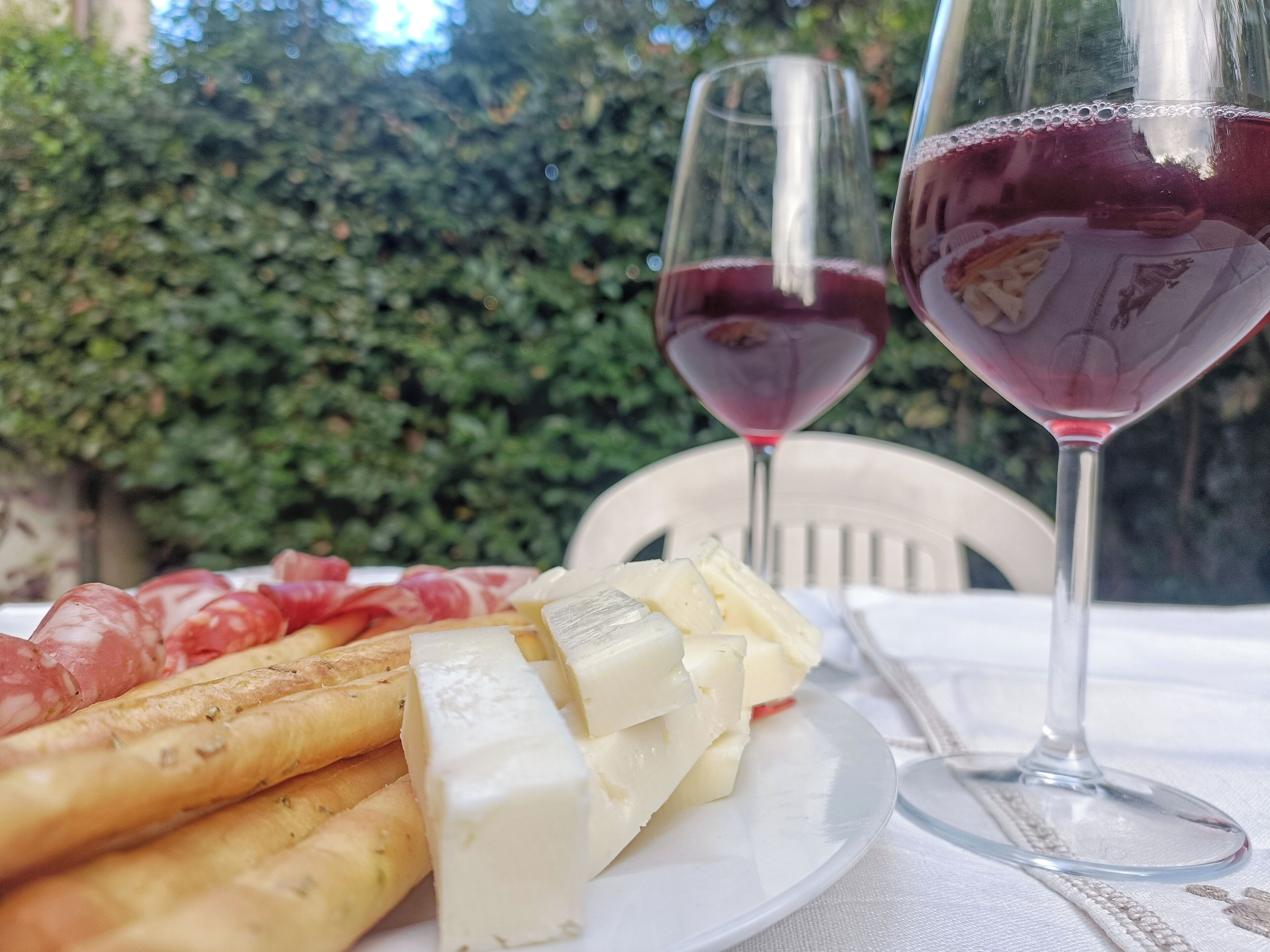 Une assiette garnie de différents fromages et de charcuteries, deux verres à vin remplis de vin rouge et une chaise blanche.