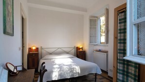 A bedroom with a bed, two bedside tables with lamps, a radiator, a window with a view, and a door with a checkered curtain. - The key to time (Lucca)