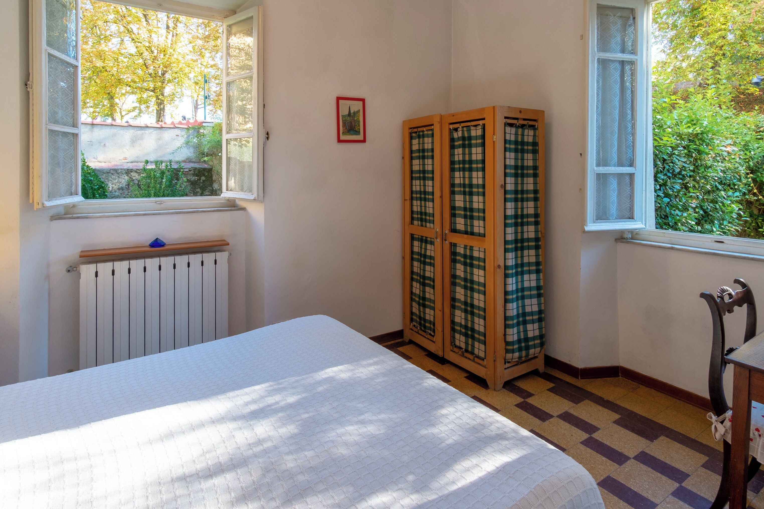 A room with a bed, a radiator, a window with a view of trees, and a checkered cabinet.