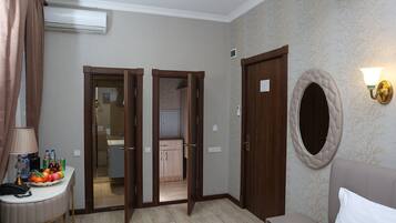 Una camera d'albergo con un letto, un comodino con una lampada, uno specchio e un punto luce a parete. Si vede una porta che conduce al bagno e a una piccola cucina.