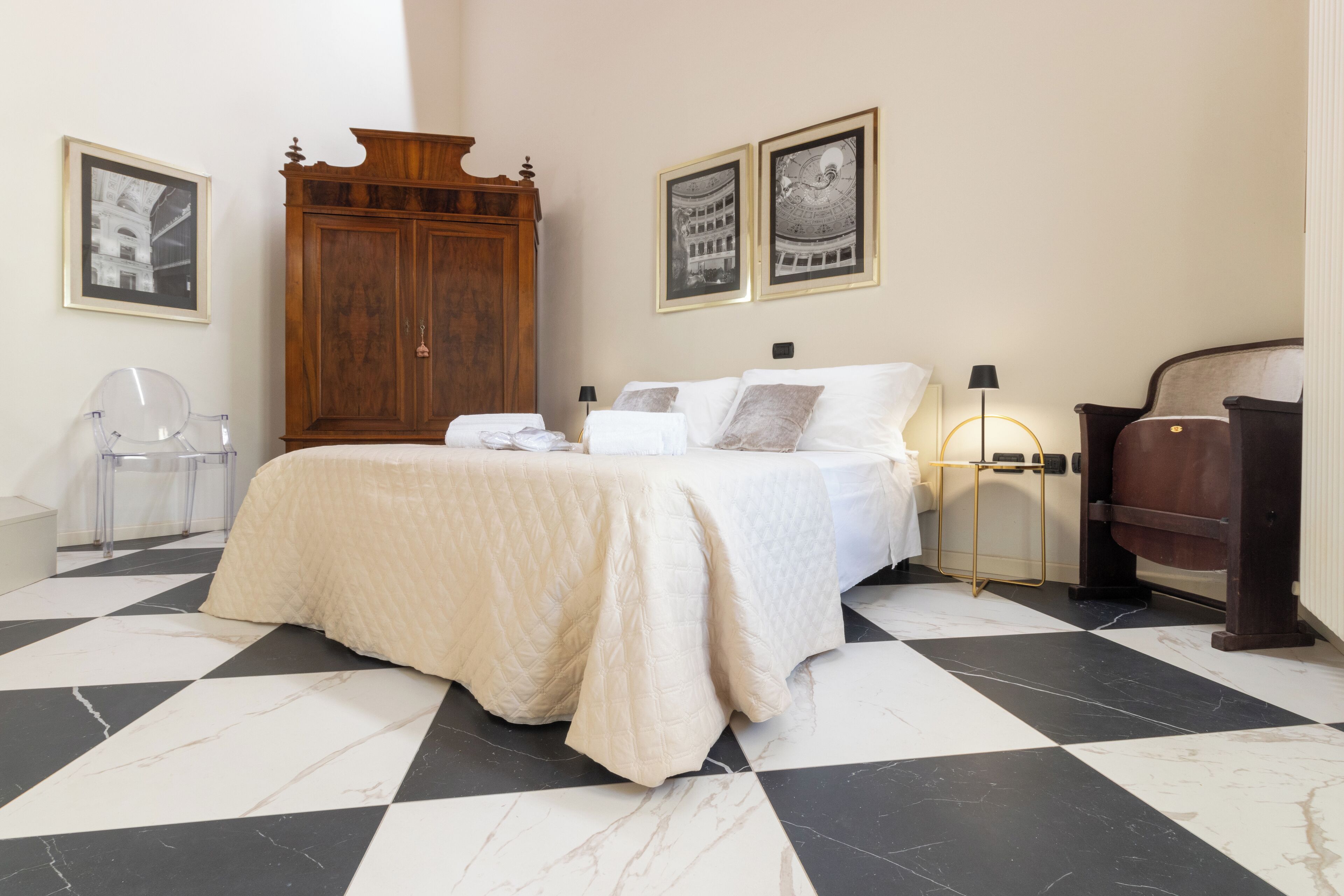 A hotel room with a checkered floor, a bed with white bedding, a wooden wardrobe, and two framed pictures on the wall.