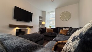 A modern living room with a large flat-screen TV mounted on the wall, a round wall clock, and a cozy sectional sofa with multiple cushions.