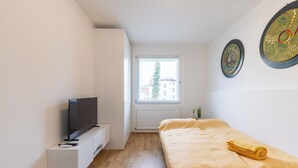 Un dormitorio moderno con una cama, un televisor sobre un mueble blanco, una ventana con vistas a edificios y dos obras de arte en la pared.
