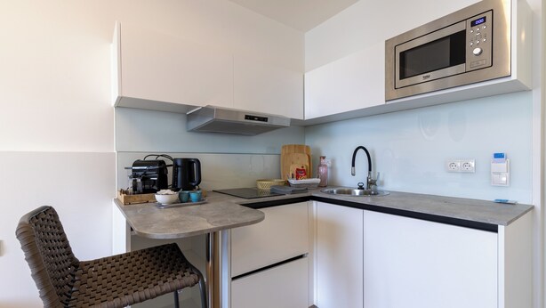 Una cocina compacta con armarios blancos, un microondas de acero inoxidable, una cafetera y un taburete de bar.