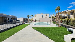 A modern outdoor pool area with a clear blue sky, surrounded by neatly trimmed grass and palm trees. - Townhouse with sea views Evergreen Homes (Mijas Costa)