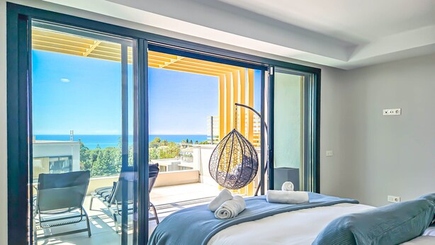 A modern bedroom with a large bed, a hanging chair, and a view of the sea. - Townhouse with sea views Evergreen Homes (Mijas Costa)