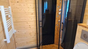 Habitación con paneles de madera, ducha con mampara de vidrio, radiador blanco y un taburete de madera.
