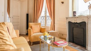 A cozy living room with a beige sofa, a matching armchair, a glass coffee table with a book and a teapot, and a fireplace.