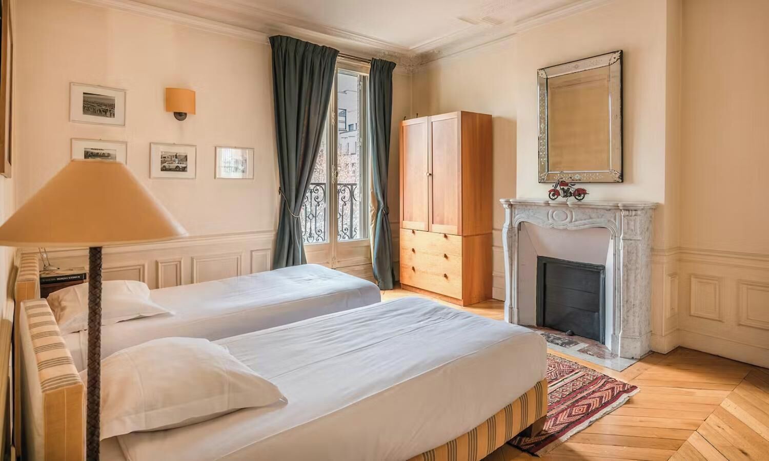 A bedroom with two beds, a wardrobe, a fireplace, and framed pictures on the wall.