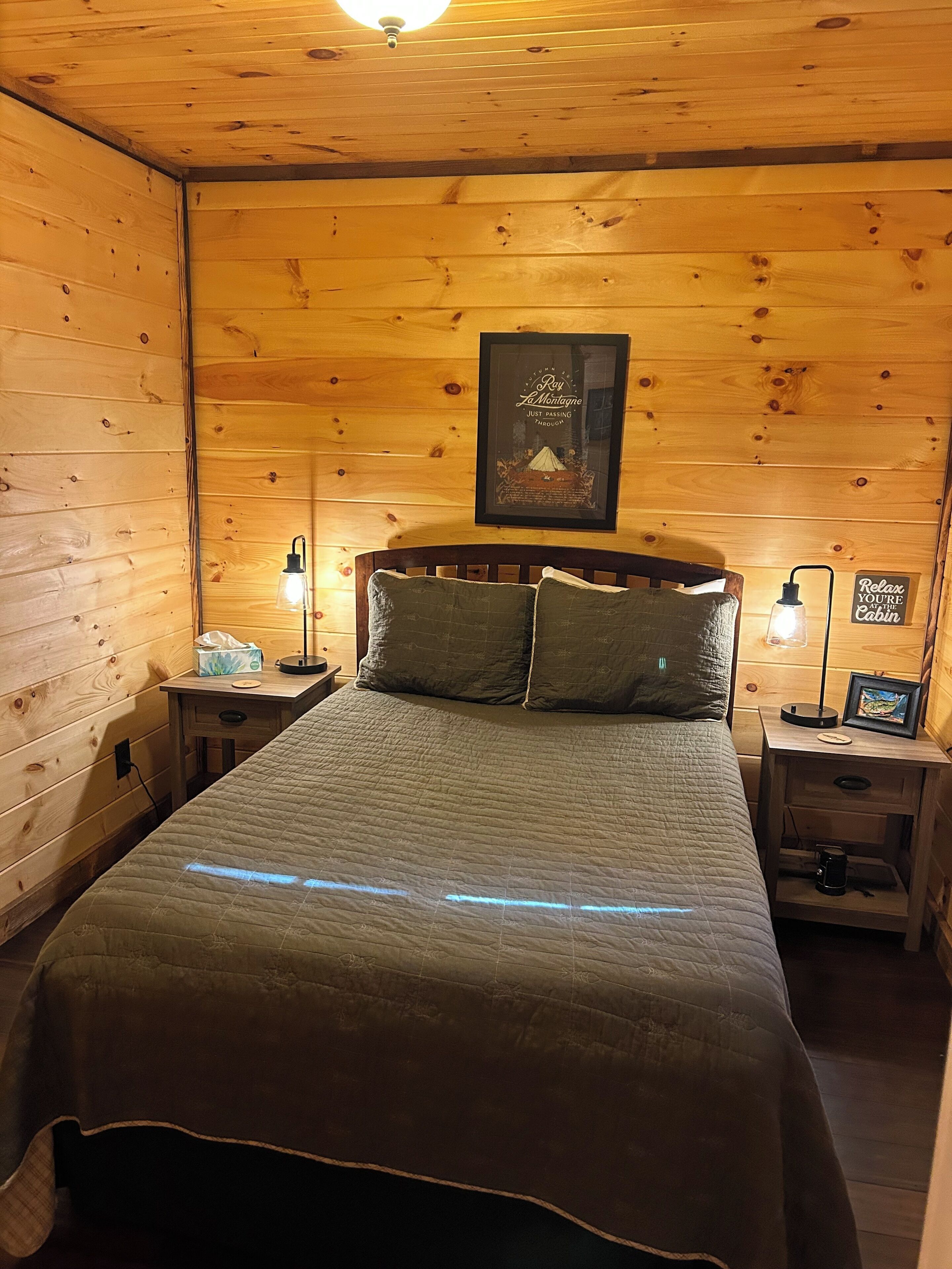 Une chambre dans un chalet en bois, avec un lit, deux tables de chevet, un tableau encadré au mur et une pancarte accrochée au mur.