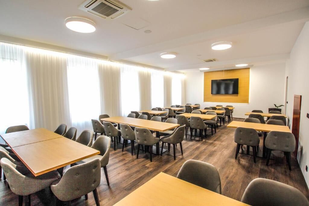 A conference room with multiple tables and chairs, large windows with curtains, a mounted flat-screen TV, and a wooden floor.