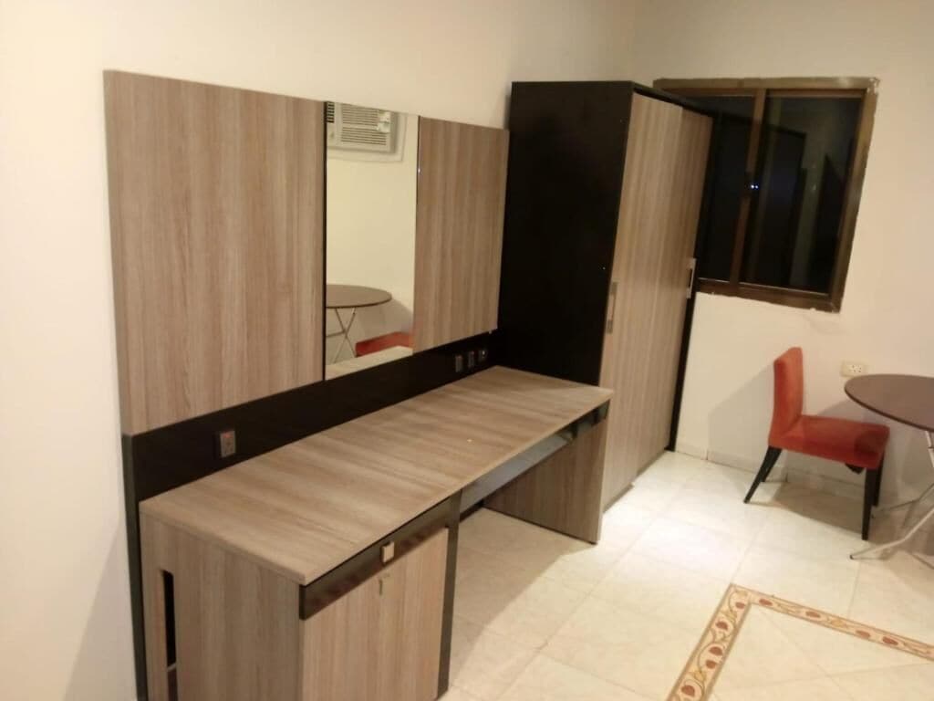 Quarto compacto com escrivaninha embutida, uma cadeira, uma mesinha e uma janela.