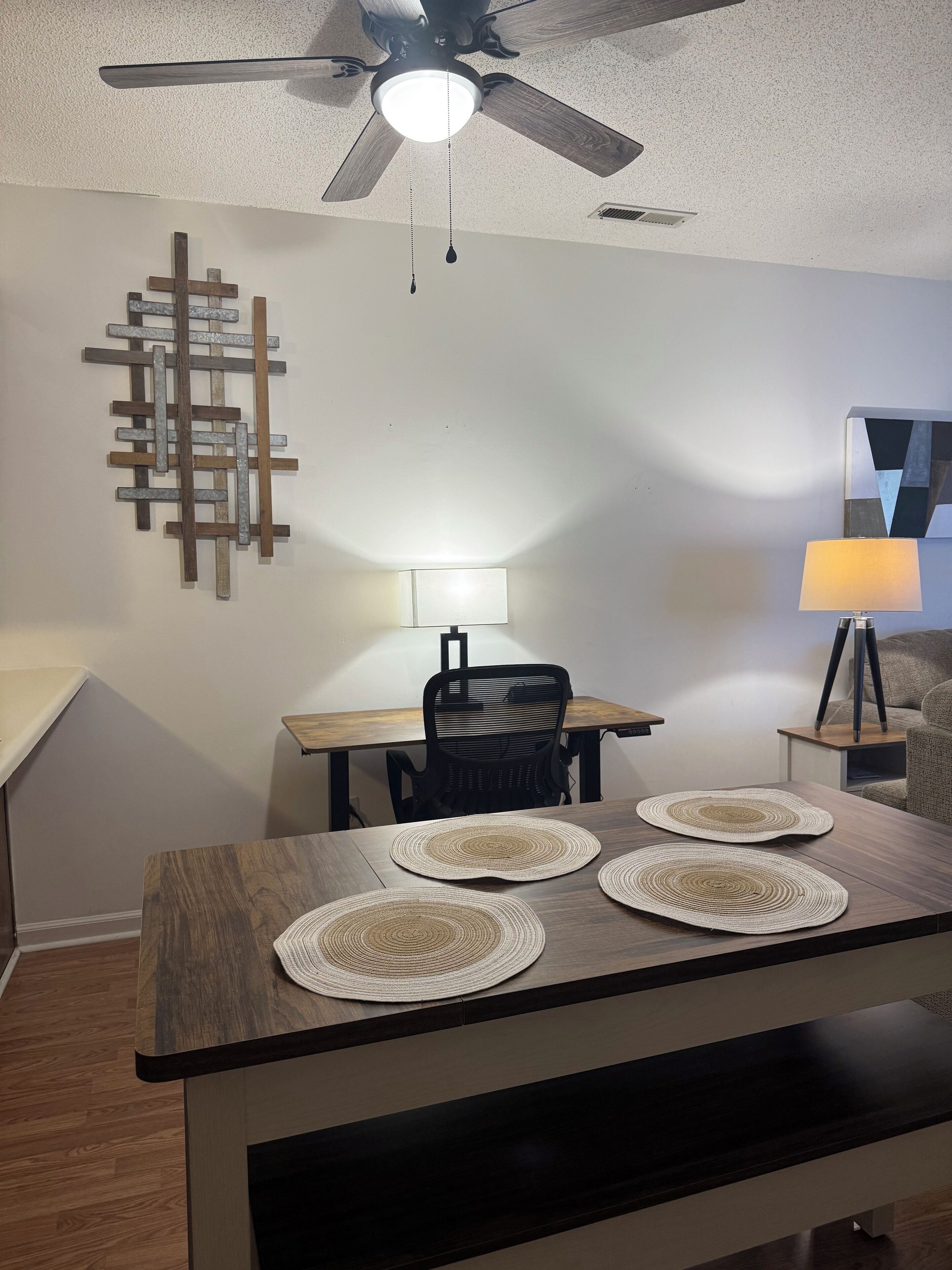A dining area with a wooden table, three woven placemats, a ceiling fan, a wall-mounted light, and a modern abstract art piece.