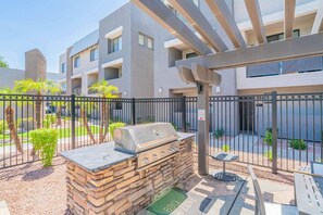 Zona de barbacoa con una parrilla de piedra, un pérgola y un espacio exterior vallado con un edificio al fondo.