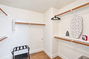 A room with a wooden floor, white walls, and a black metal chair. There are two wooden beams on the wall, one with a white towel hanging and another with a black hanger. A white shelf with a hexagonal mirror and a small iron is also visible. - Walk To Westgate, Pool, Gym, WD & Free Parking (Glendale)