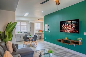 A modern living room with a television, a dining area, and a kitchen in the background. - Walk To Westgate, Pool, Gym, WD & Free Parking (Glendale)